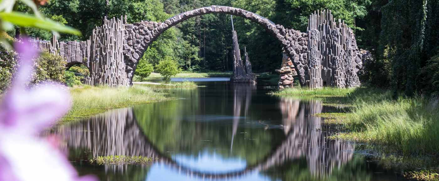 Steinbogenbrücke über Fluss mit Basaltsteinen, umgeben von grüner Natur und lila Blüten im Vordergrund