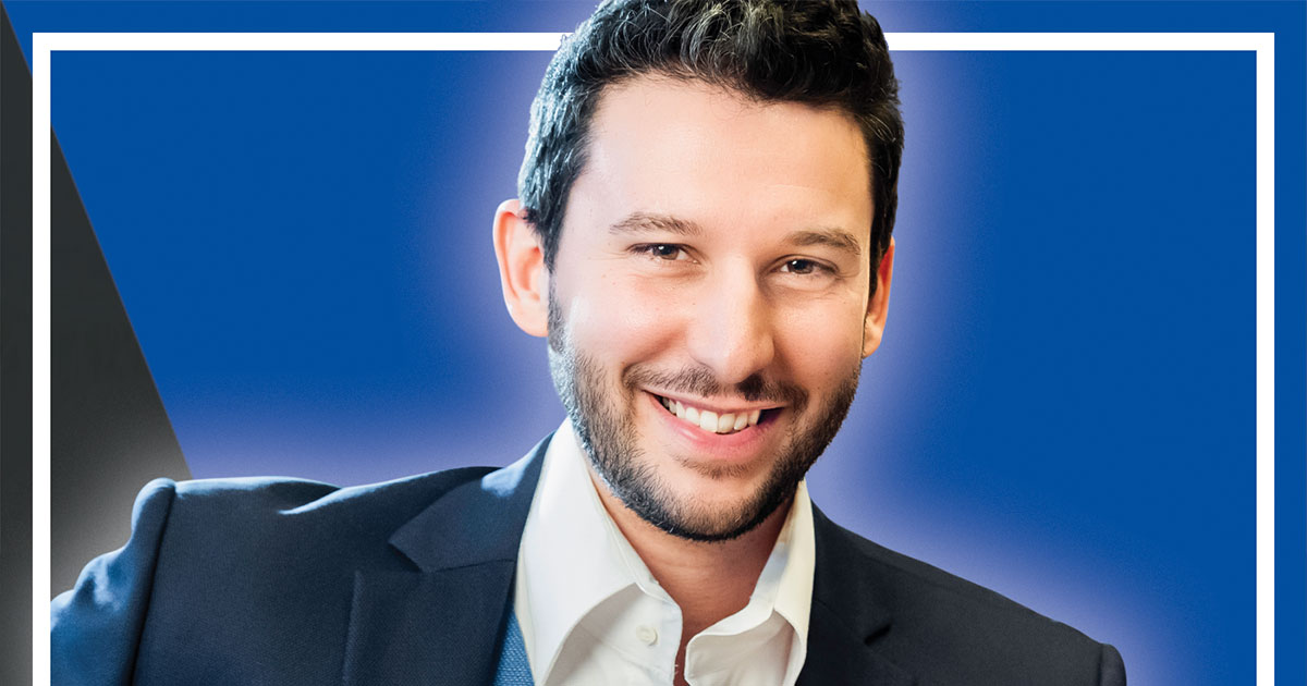 Benedikt Ahlfeld lächelnd im Business-Anzug vor blauem Hintergrund mit weißem Rahmen