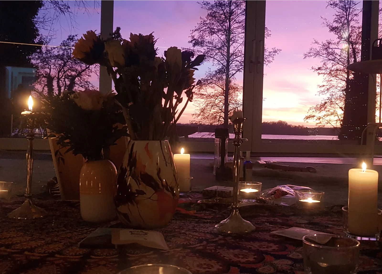 Sonnenuntergang über See mit Blick auf getrocknete Sonnenblumen, Kerzen und Altar vor Fenster