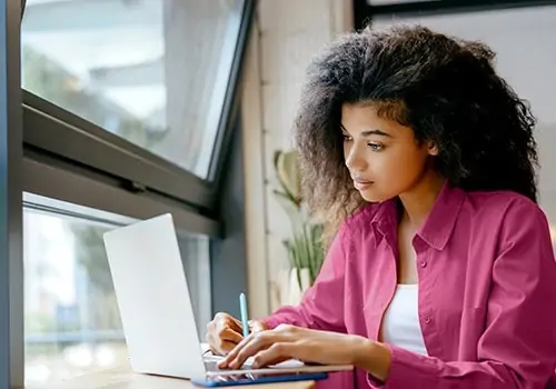 Frau in rosa Hemd arbeitet am Laptop am Fenster, Online-Weiterbildung