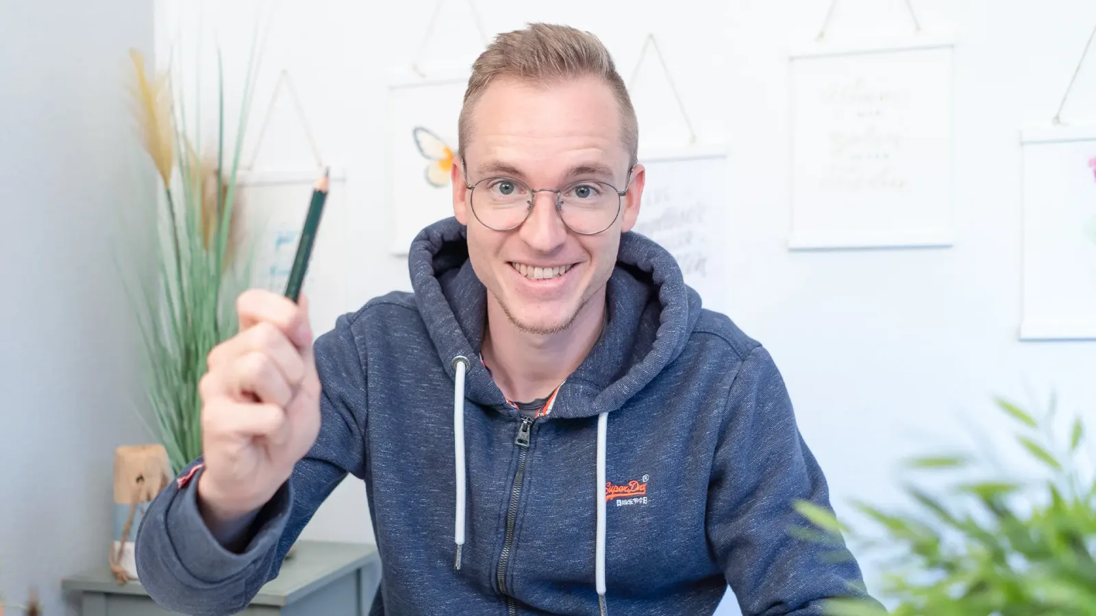 Mann mit Brille und Stift zeigt Spiralblock mit Banner-Skizzen, lächelt zur Kamera