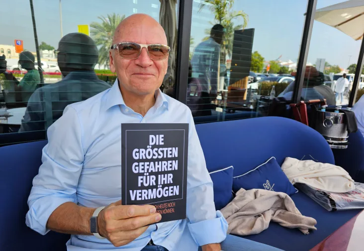 Mann mit Brille hält Buch "Die größten Gefahren für Ihr Vermögen" in modernem Restaurant