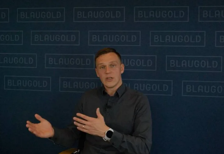 Mann in schwarzem Hemd vor BLAUGOLD-Hintergrund sprechend