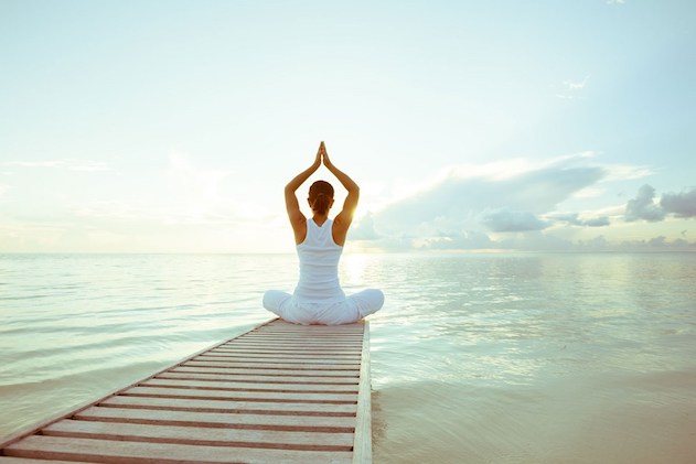 Frau in weißem Top sitzt in Yoga-Pose auf Steg über ruhigem Meer bei Sonnenaufgang