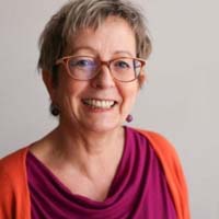 Portraitfoto: Frau mit rötlichem Haar, Brille und farbigem Top auf neutralem Hintergrund, lächelnd