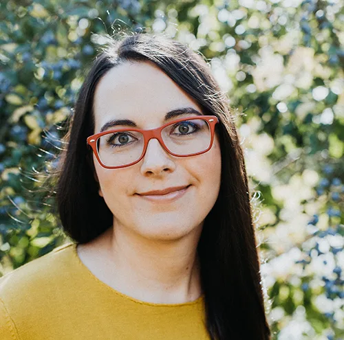 Portraitfoto: Junge Frau mit schwarzen Haaren, roter Brille und gelbem Oberteil vor grünem Hintergrund