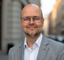 Portrait-Foto von Christian Költringer in grauem Sakko und weißem Hemd vor städtischem Hintergrund