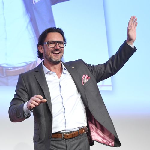 Oliver Geisselhart auf der Bühne mit Brille, grauem Blazer und weißem Hemd, winkt dem Publikum zu