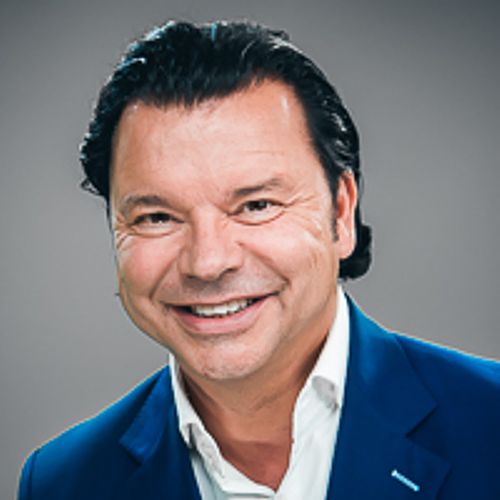 Paul Misar mit dunklem Anzug und blauem Hemd, freundlich lächelnd auf grauem Hintergrund