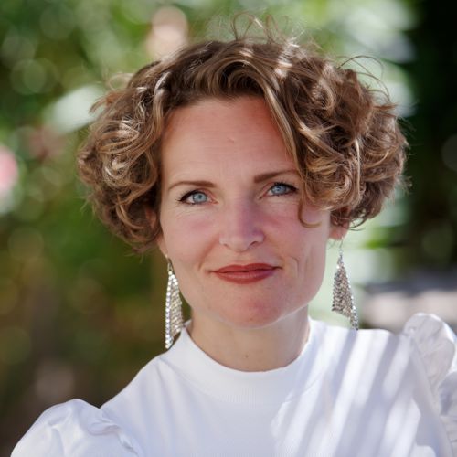 Stefanie Bruns mit lockigem braunem Haar, weißem Oberteil und Perlenohrring vor grünem Hintergrund