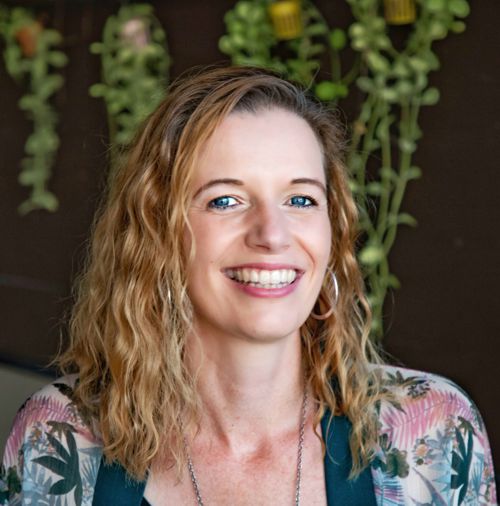 Tanja Baumann mit welligem rotblondem Haar vor grünem Pflanzenhintergrund, strahlendes Lächeln