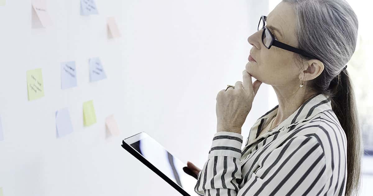 Frau mit Brille und gestreiftem Oberteil hält Tablet, schaut auf Notizen an der Wand
