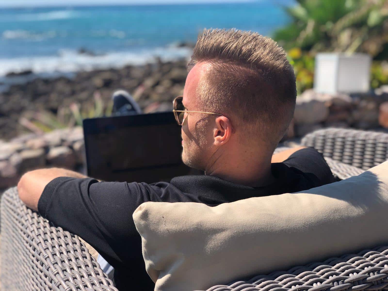 Mann mit Brille sitzt am Strand mit Laptop, Meer im Hintergrund, arbeitet ortsunabhängig