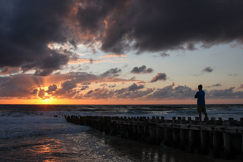 Mann im Profil auf Wellenbrecher bei Sonnenuntergang mit rotem Himmel