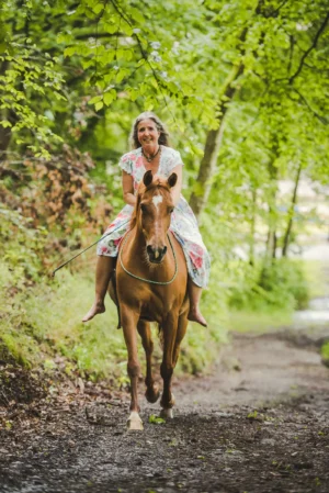 Christiane Göbel reitet auf braunem Pferd durch grüne Baumgasse, trägt geblümtes Kleid