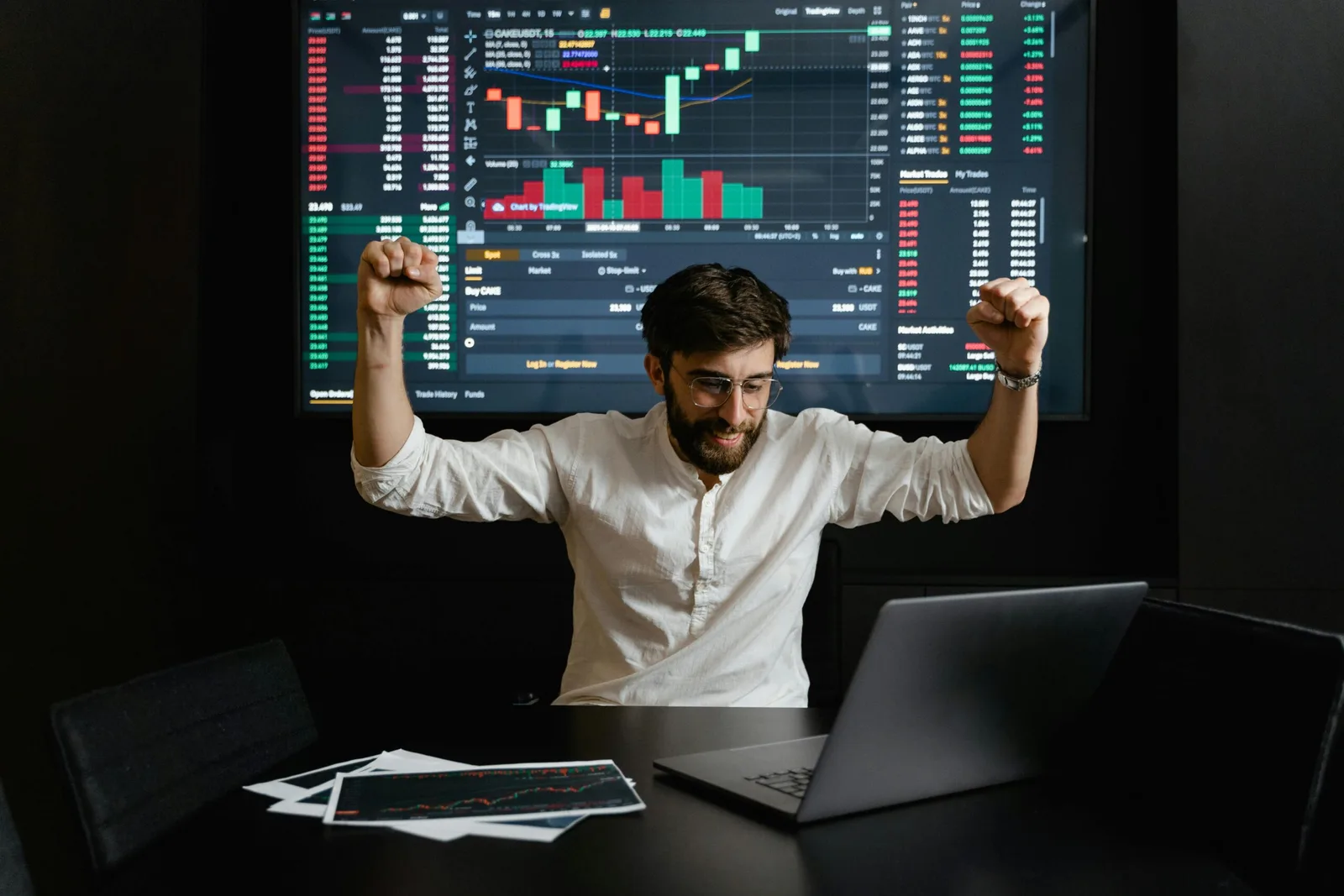 Mann am Schreibtisch mit erhobenen Armen vor großem Finanz-Dashboard mit Graphen
