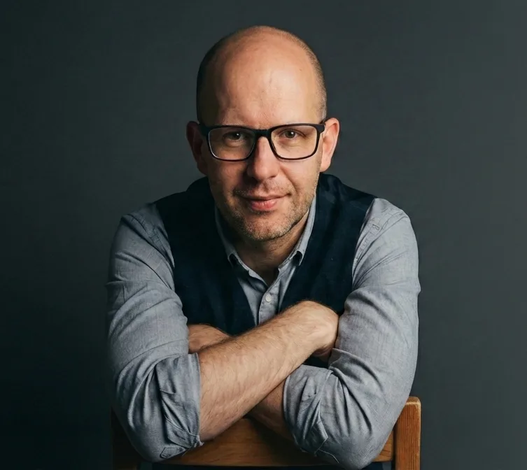 Porträt von Michael Schlinder mit Brille, grauem Hemd und dunkelblauem Weste vor grauem Hintergrund