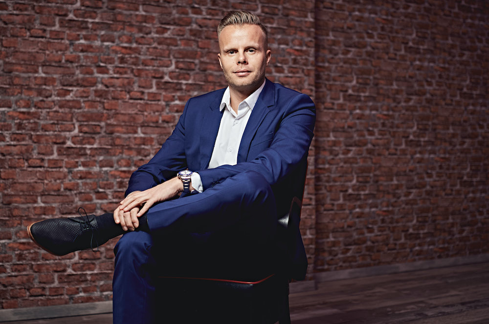 Gunnar Kessler in dunkelblauen Anzug sitzt auf Stuhl vor Backsteinwand