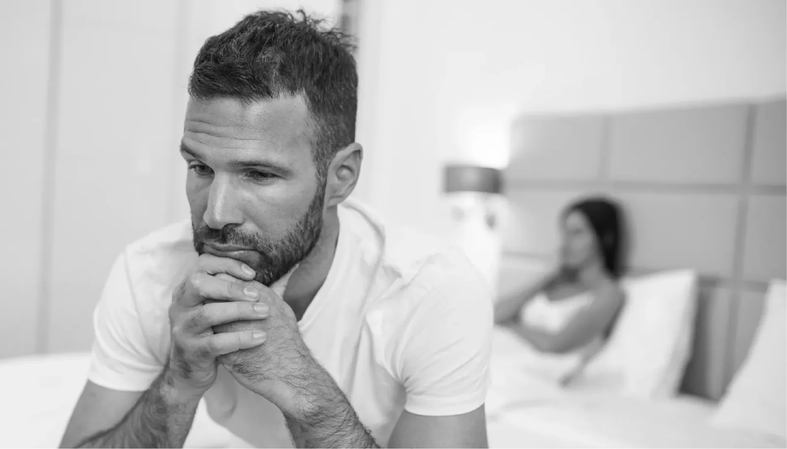 Schwarzweiß-Fotografie eines nachdenklichen Mannes in weißem T-Shirt mit verschwommener Frau im Hintergrund