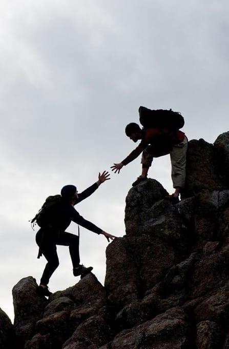 Person hilft anderer Person, Felsen hochzuklettern, Silhouette gegen bewölkten Himmel