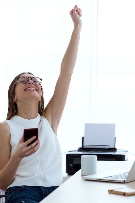 Frau am Schreibtisch mit erhobener Faust, Smartphone in Hand, Laptop und Büromaterialien im Hintergrund