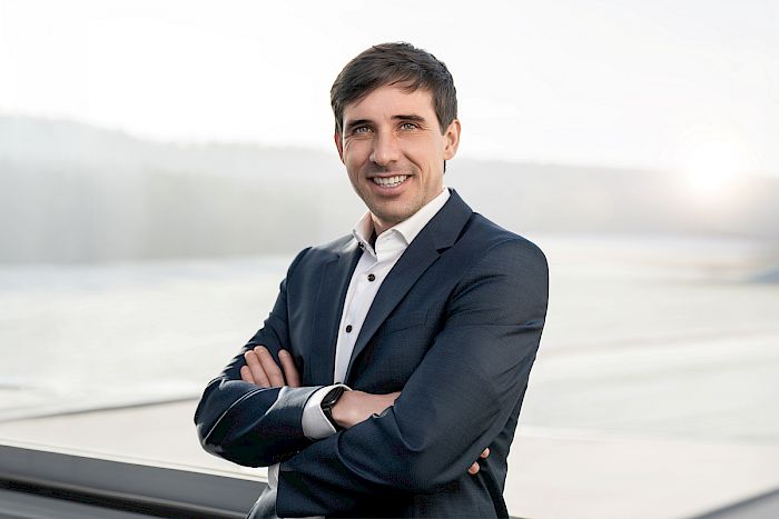 Porträt eines Mannes im dunkelblauen Anzug vor Fenster mit Seenlandschaft