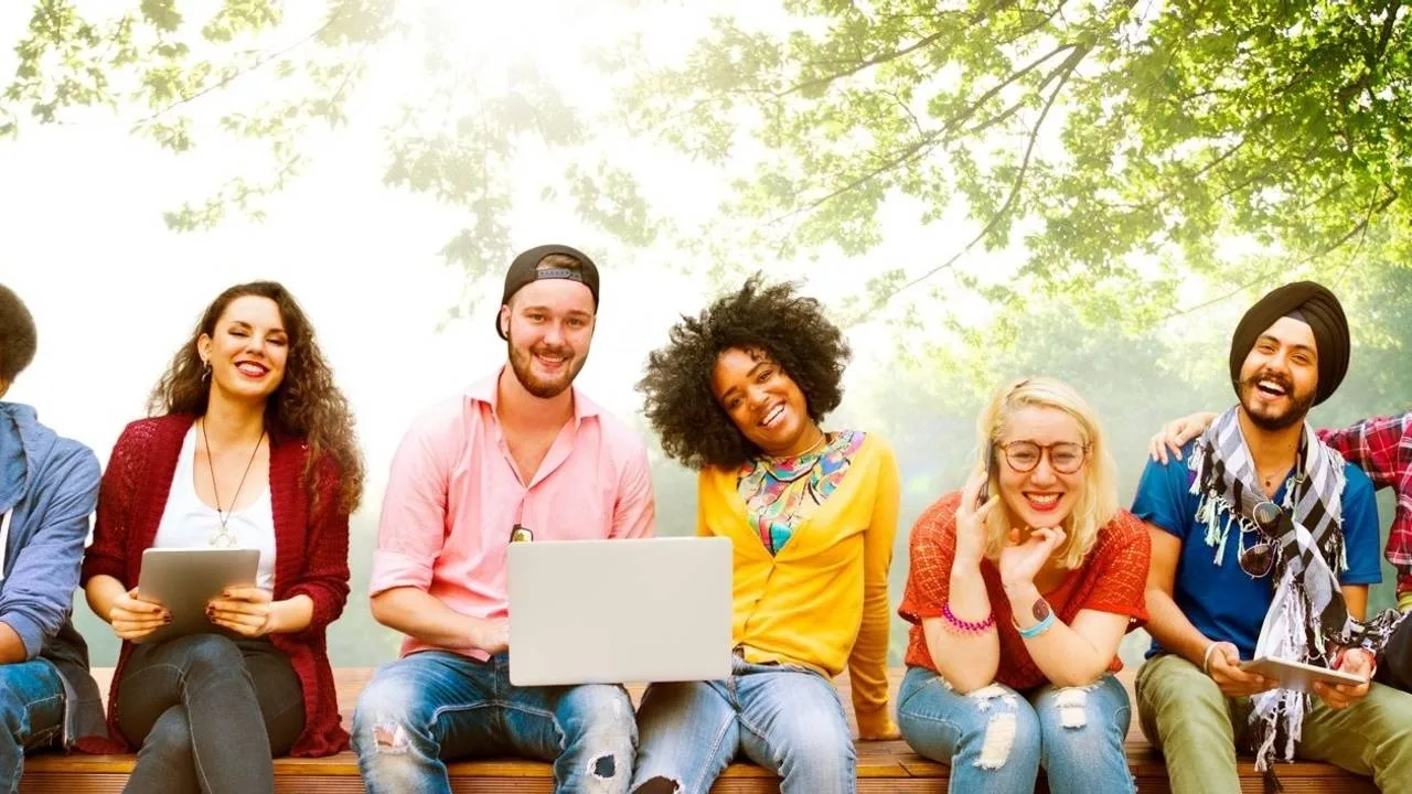 Diverse Gruppe junger Menschen sitzt auf Bank mit Laptop und Tablet unter grünen Bäumen
