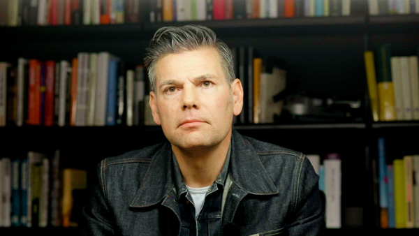 Porträt Ken Jebsen vor Bücherregal, graues Hemd, ernsthafter Blick