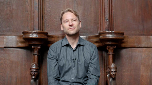 Porträt Konstantin Wolff vor geschnitztem Holzpaneeel, graues Hemd, Mikrofon
