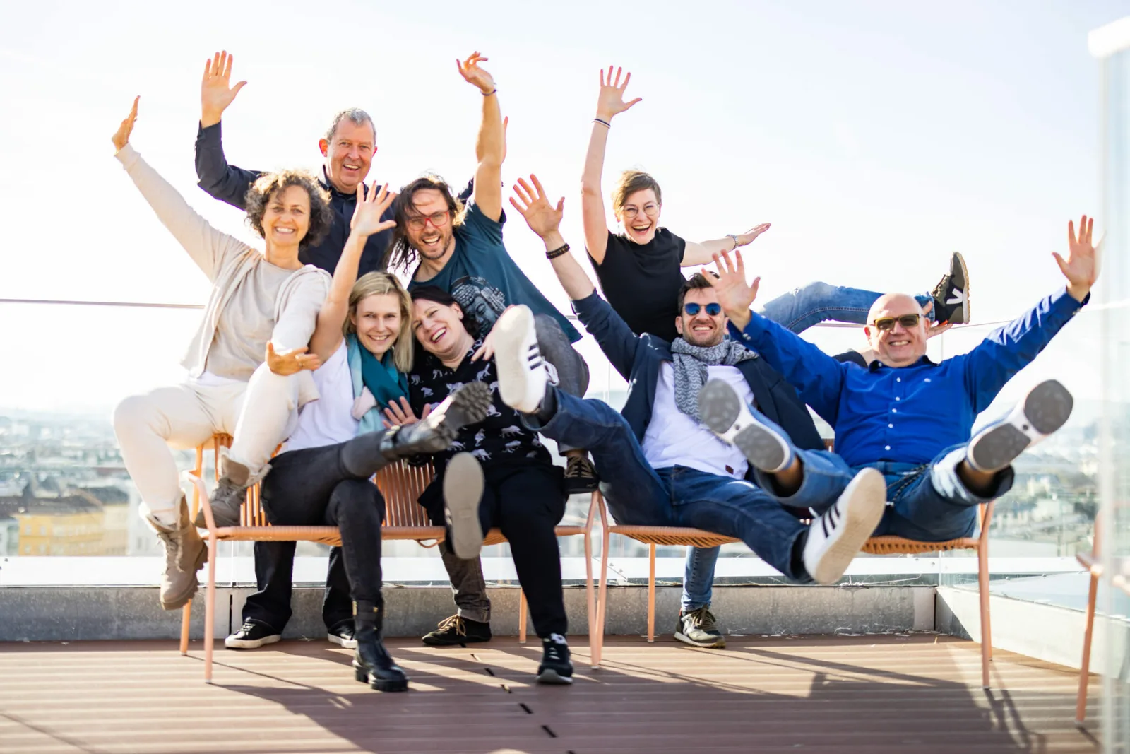 Gruppe von zehn jubelnd Personen auf Dachterrasse mit Stadtskyline, Arme hochgestreckt