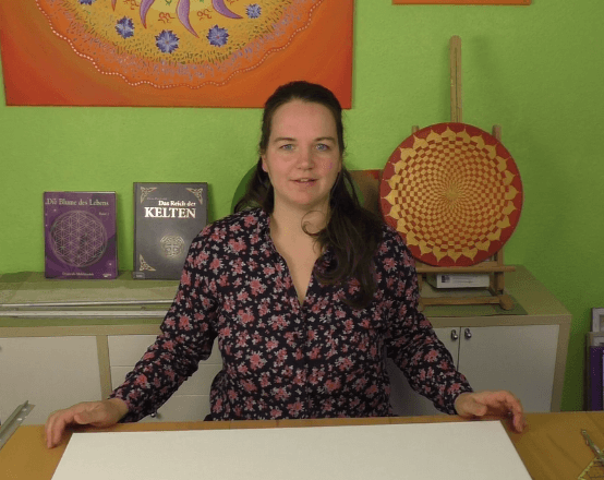 Energiebilder selber malen – Online Videomalkurs Mandala 31 Frau vor grüner Wand mit Mandala-Kunstwerken und Büchern im Hintergrund