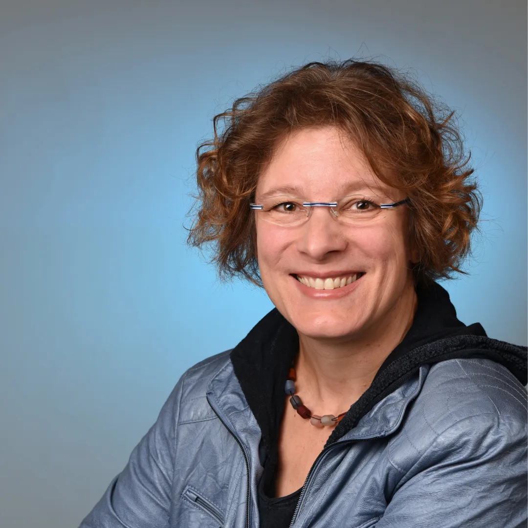 Porträt Corinna mit rötlichbraunem lockigem Haar und Brille auf blauem Hintergrund