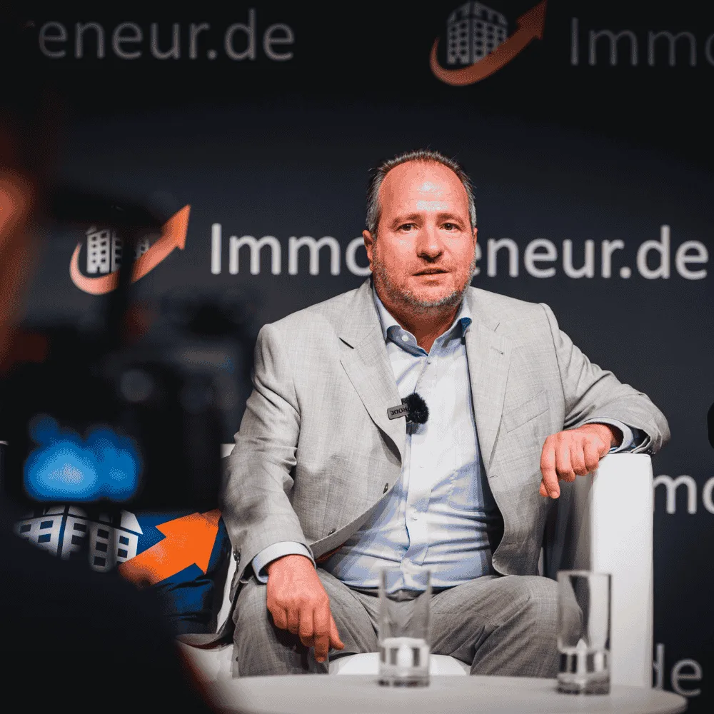 Thomas Knedel im Interview vor Immopreneur.de Logo, beige Anzug mit Mikrofon