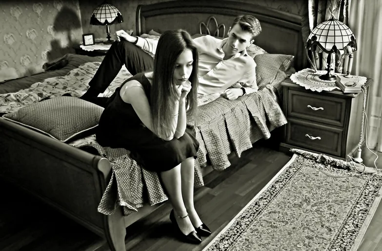 Schwarz-Weiß-Fotografie einer Frau auf einem Bett mit einem Mann im Hintergrund, Schlafzimmer-Szenario