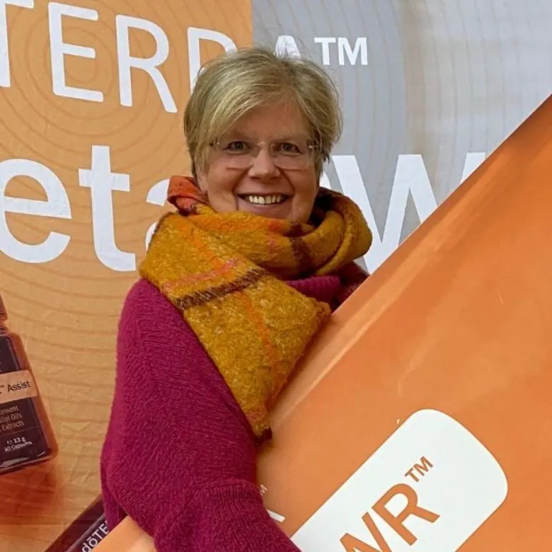 Ernährungsberatung – Coaching für anti-entzündliche Ernährung 7 Nicola Herrmann mit Schal vor Logo-Wand bei Konferenz, hält Produktbox