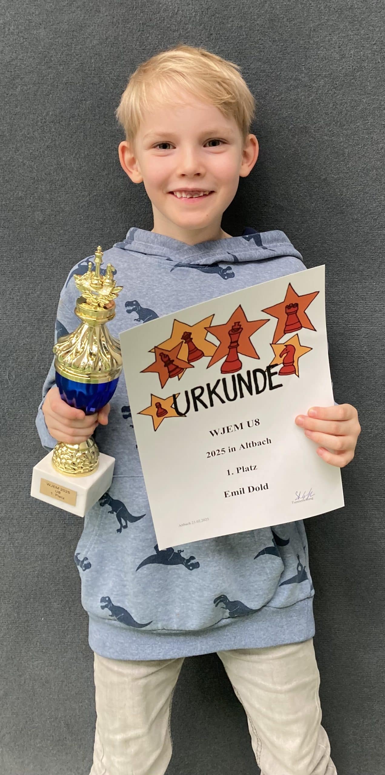 Blondes Kind mit grauem Hoodie hält Schach-Urkunde und goldene Trophäe lächelnd
