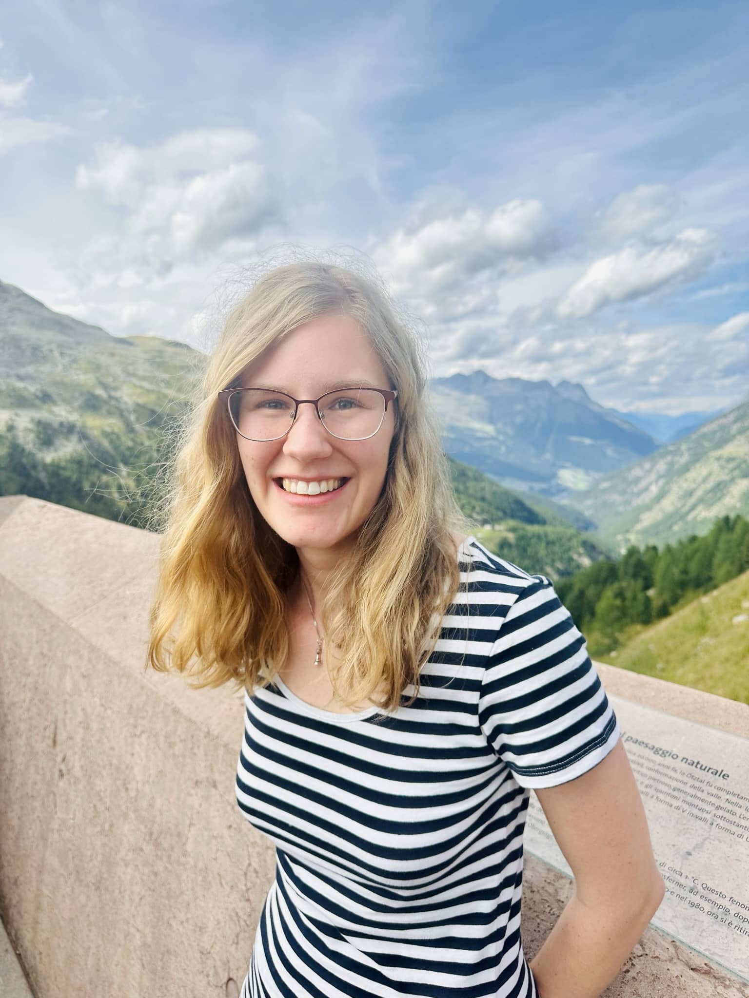Junge Frau mit Brille lächelt vor Berglandschaft in gestreiftem Shirt
