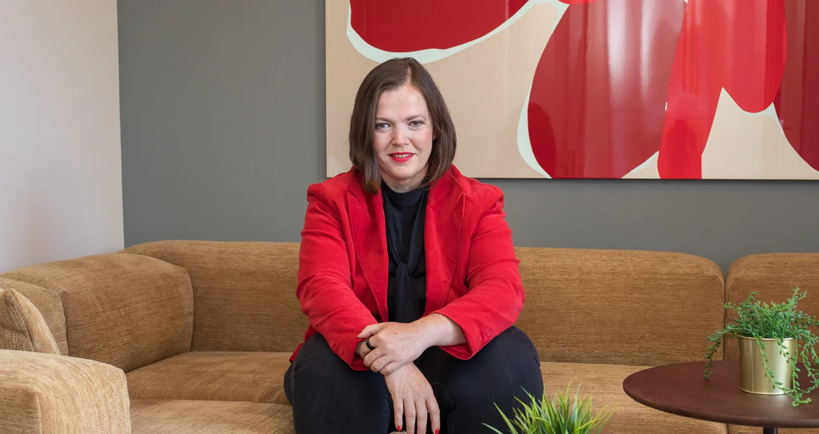 Petra Binder in rotem Blazer auf beigem Sofa sitzt – modernes Büro-Setting