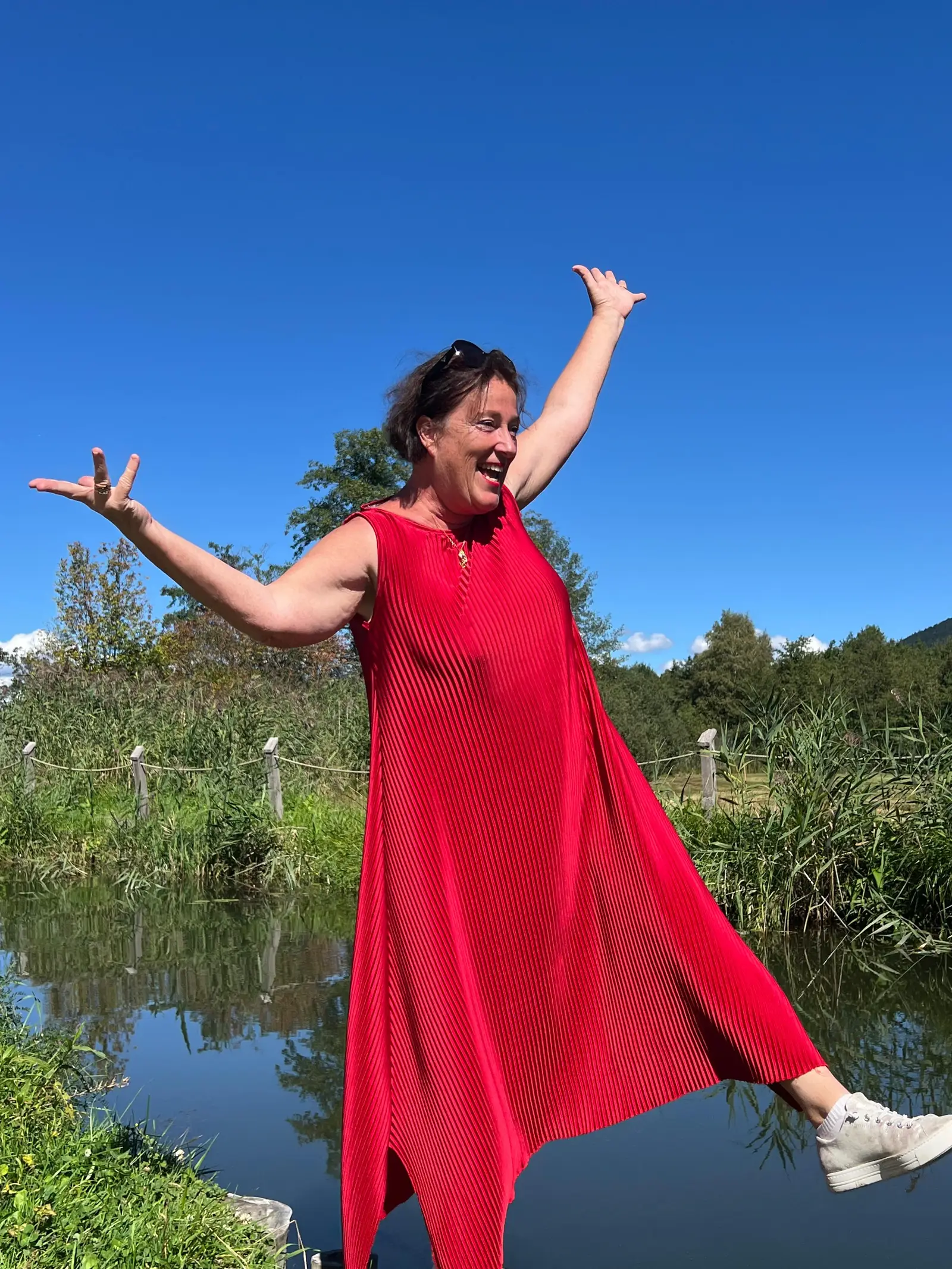 Frau in rotem Kleid mit ausgestreckten Armen an Teich und Wald, Freude und Leichtigkeit