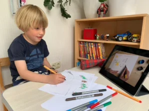Junge zeichnet mit Buntstiften auf Papier, auf Tablet läuft Video-Anleitung