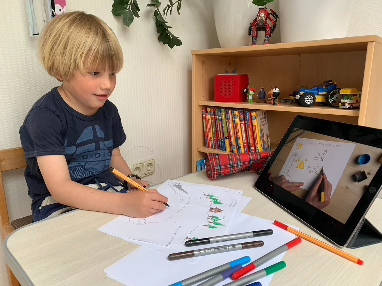 Junge zeichnet mit Buntstiften auf Papier, auf Tablet läuft Video-Anleitung