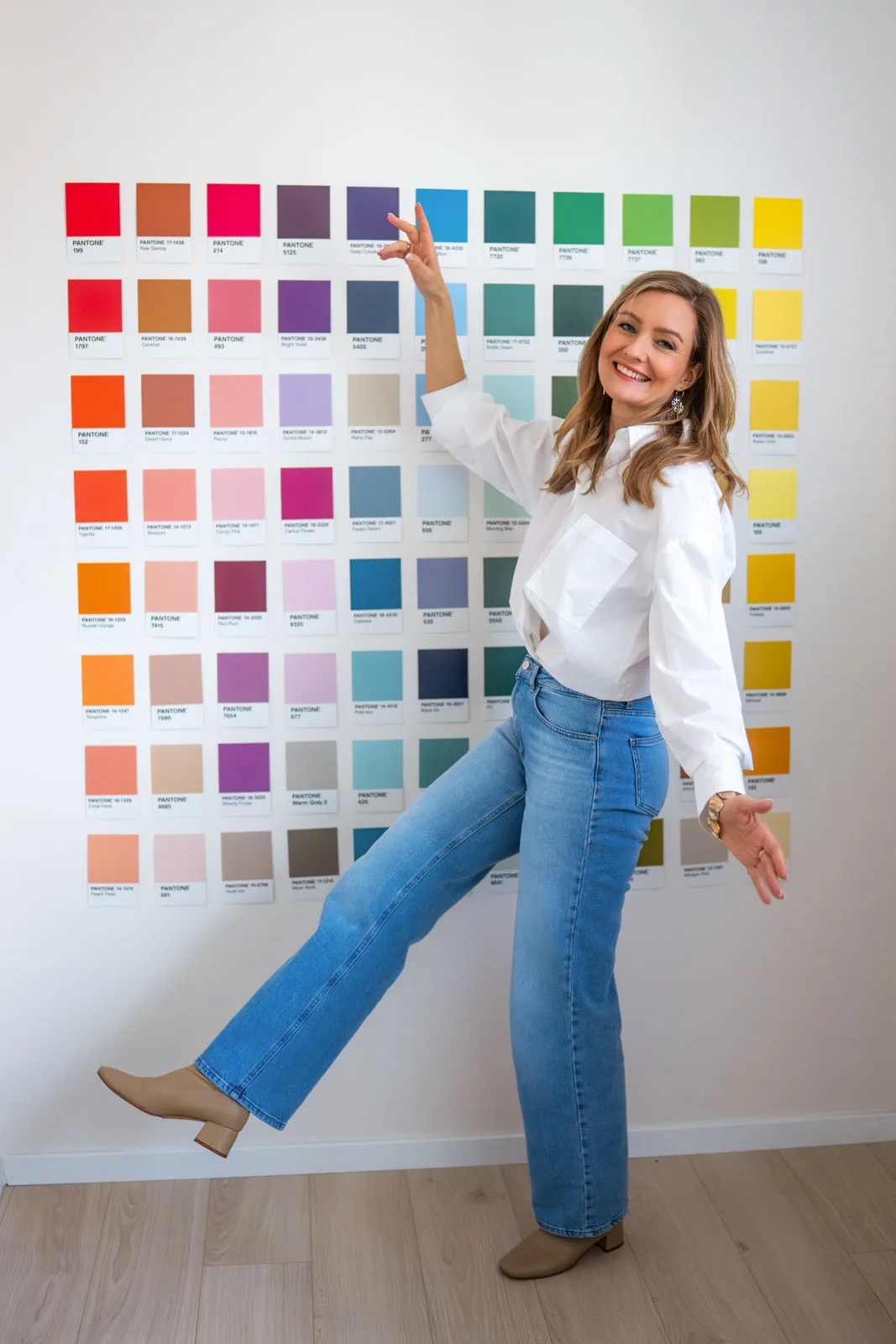 Farbe – Live-Workshop zur Farbberatung 3 Frau in weißer Bluse und Jeans vor großer Pantone-Farbpalette an Wand, zeigt auf Farbe