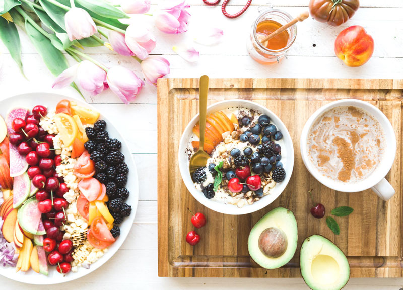 Frühstückschale mit Obst, Beeren, Avocado, Granola und Smoothie auf Holzbrett mit Blumen