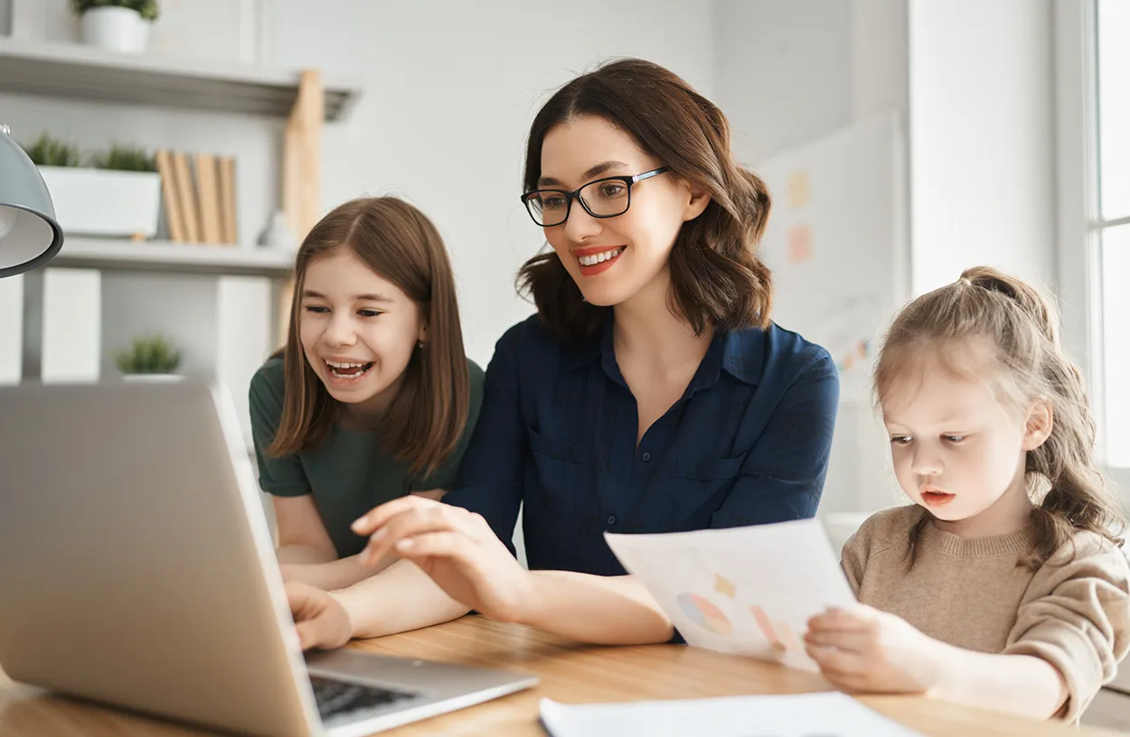 Frau mit Brille arbeitet lächelnd am Laptop mit zwei Töchtern, perfekte Balance zwischen Arbeit und Familie