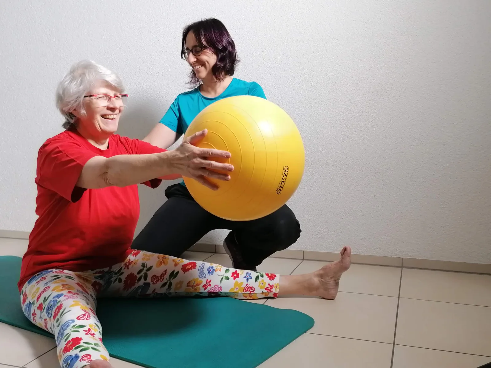 Trainerin in türkisem Shirt hilft Seniorin in rotem Shirt mit gelbem Gymnastikball, beide lächeln
