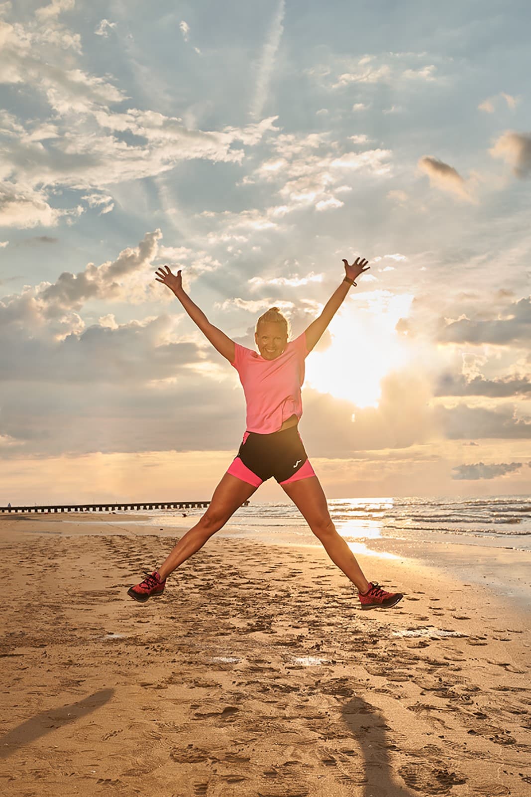 Béatrice Drach in rosa Sportoutfit springt mit erhobenen Armen am Strand vor Himmel und Meer