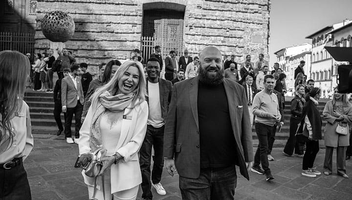Zwei Personen lachen zusammen auf historischem Platz vor Steingebäuden, Menschen im Hintergrund