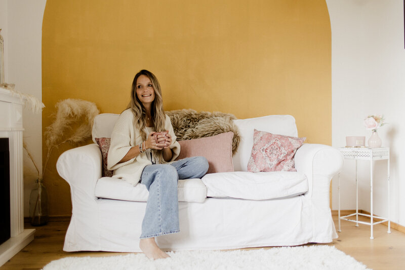Folge deinem Herzen – Community für Intuition von Sarah Jane Rogalski 3 Sarah Jane Rogalski sitzt auf weißem Sofa mit gelbem Hintergrund, trägt beiges Cardigan
