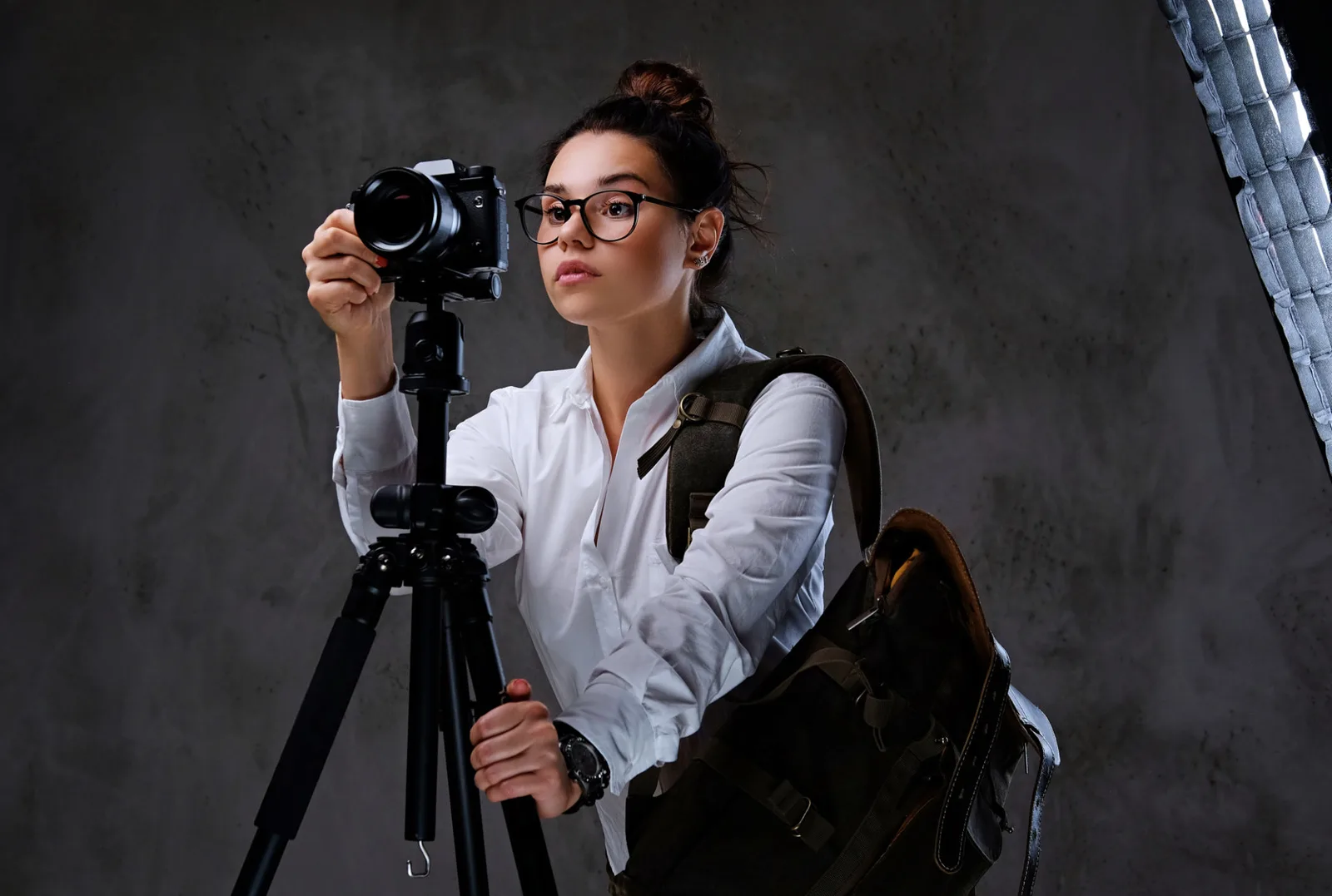 Junge Fotografin mit Brille in Weiß-Grau-Outfit mit Kamera auf Stativ vor grauem Hintergrund