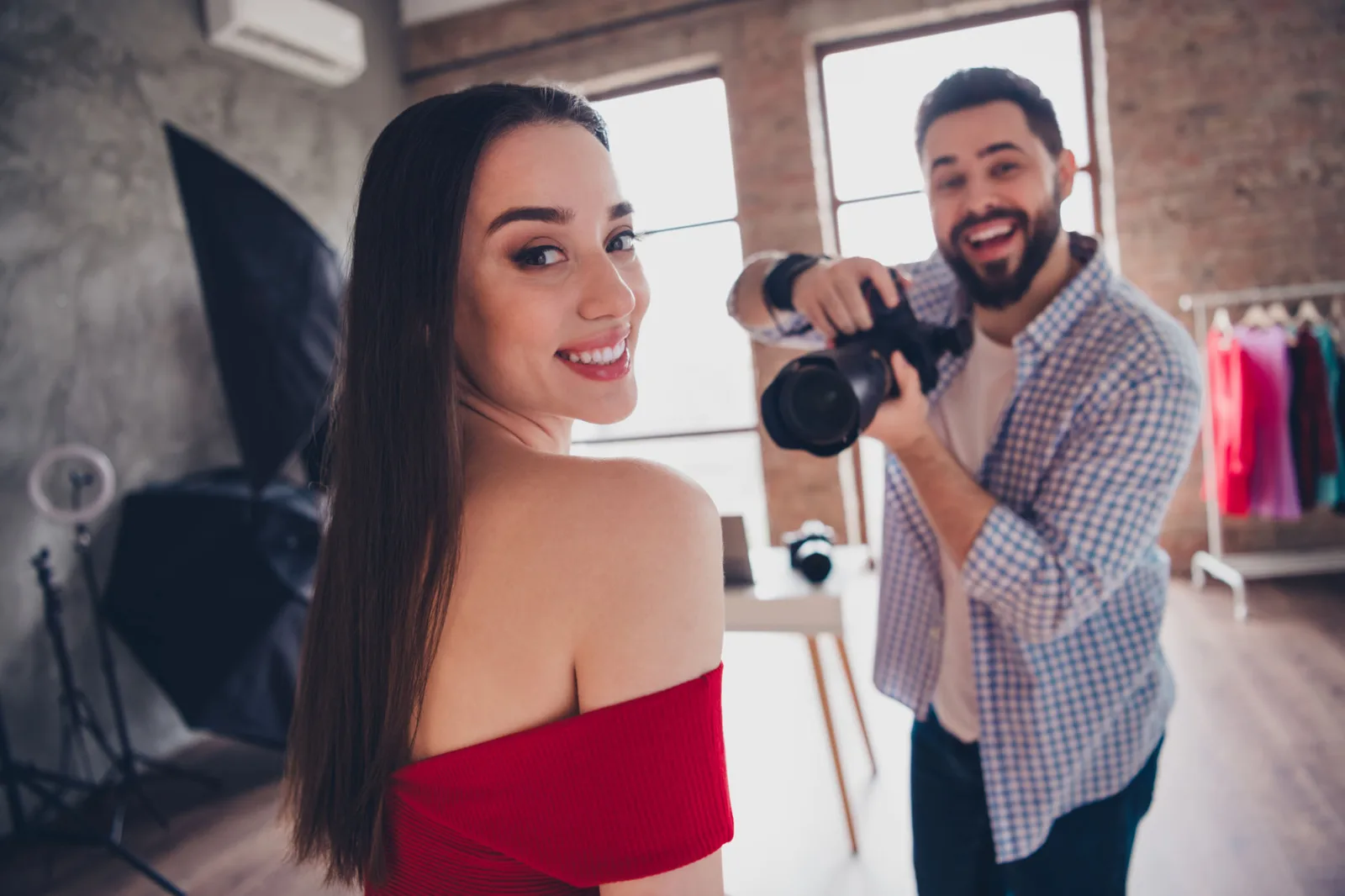 Portrait-Shooting in Studio: Fotografin posiert vor Kamera, Mann fotografiert sie, bunte Kleider im Hintergrund